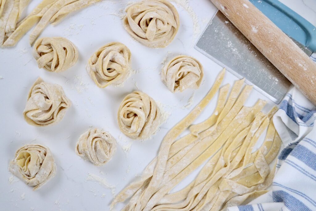 Fresh pasta being cut into fettuccine noodles by hand on a floured countertop, from scratch at home.